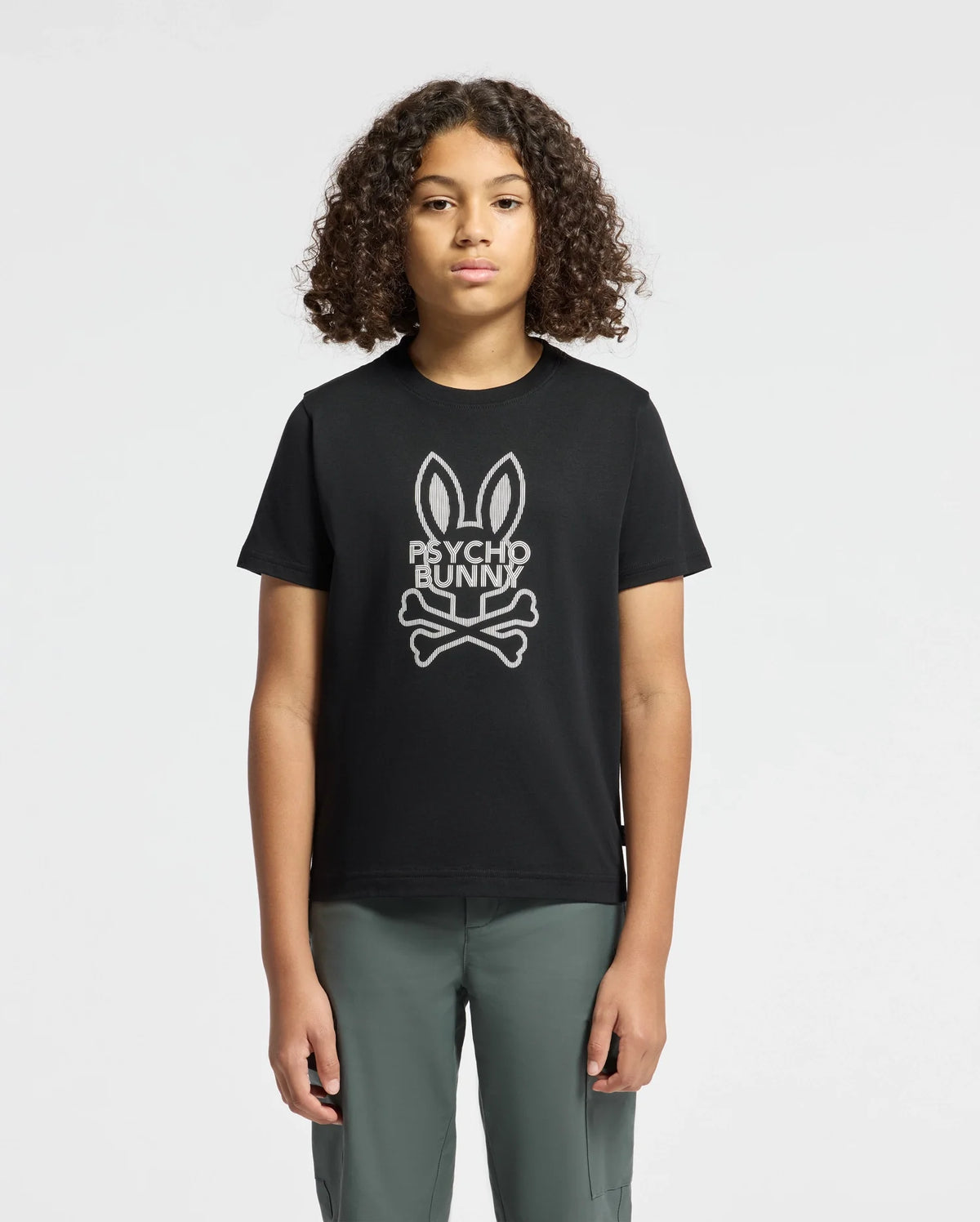 A young person with curly hair sports the KIDS DUMON GRAPHIC TEE - B0U271E200 by Psycho Bunny, showcasing a trendy bunny and crossbones design. This black T-shirt, crafted from soft Pima cotton, is elegantly paired with their gray pants against a plain white backdrop.