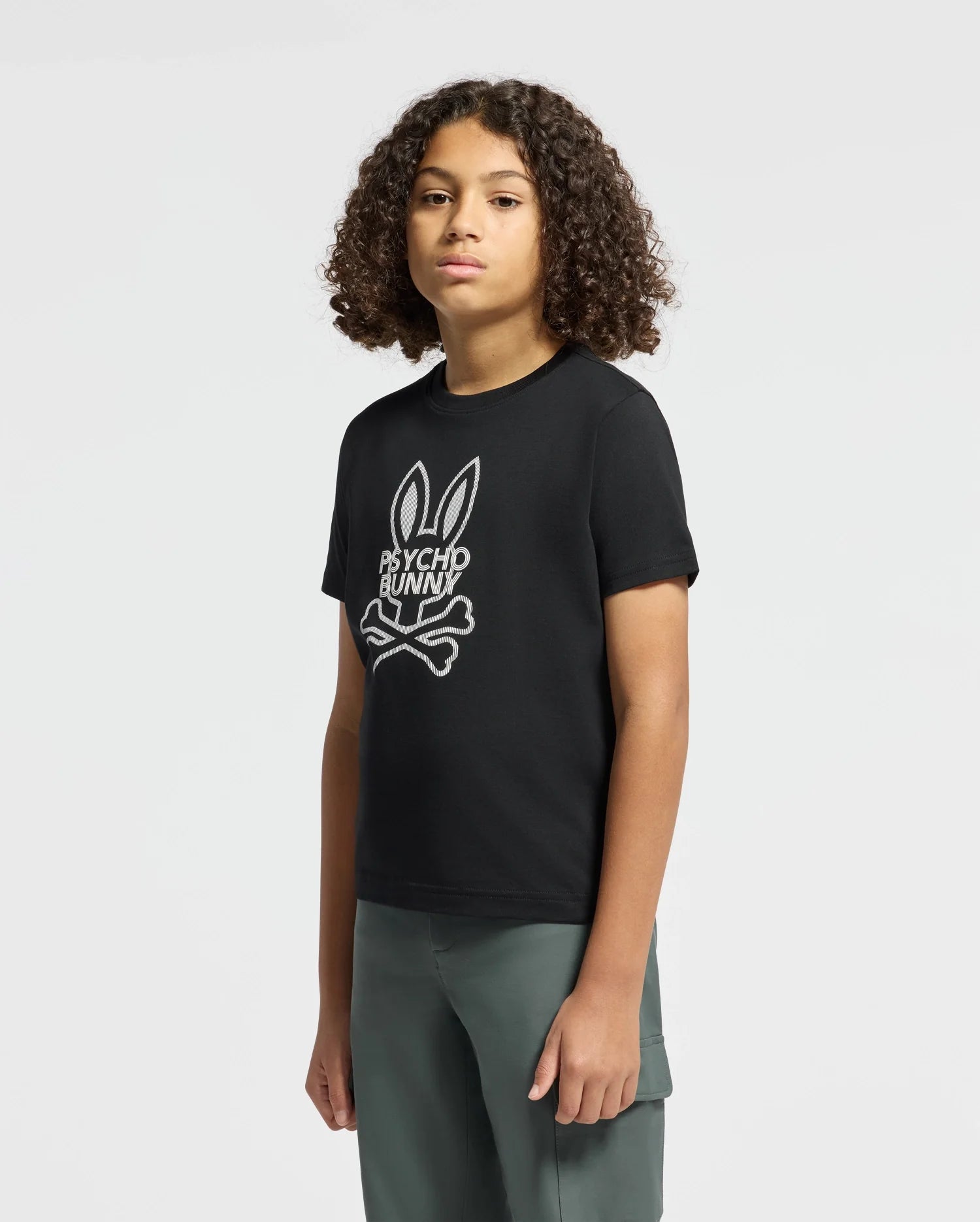 A young person with curly hair rocks the stylish KIDS DUMON GRAPHIC TEE by Psycho Bunny—an eye-catching black Pima cotton shirt featuring a distinctive white bunny head with crossbones and bold "Psycho Bunny" lettering. They stand against a plain white background, pairing the tee with green pants for an effortlessly cool look.