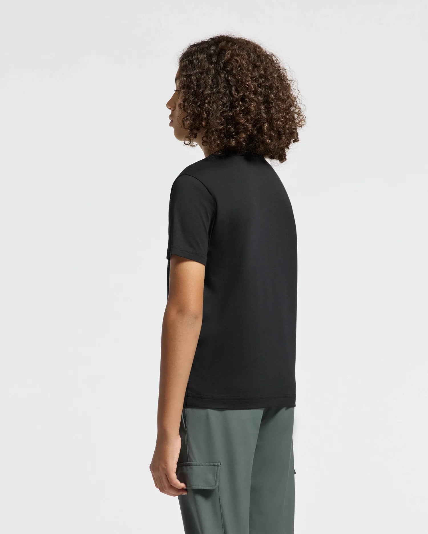 A person with curly hair stands sideways against a plain white background, wearing the stylish KIDS DUMON GRAPHIC TEE by Psycho Bunny and gray-green pants.