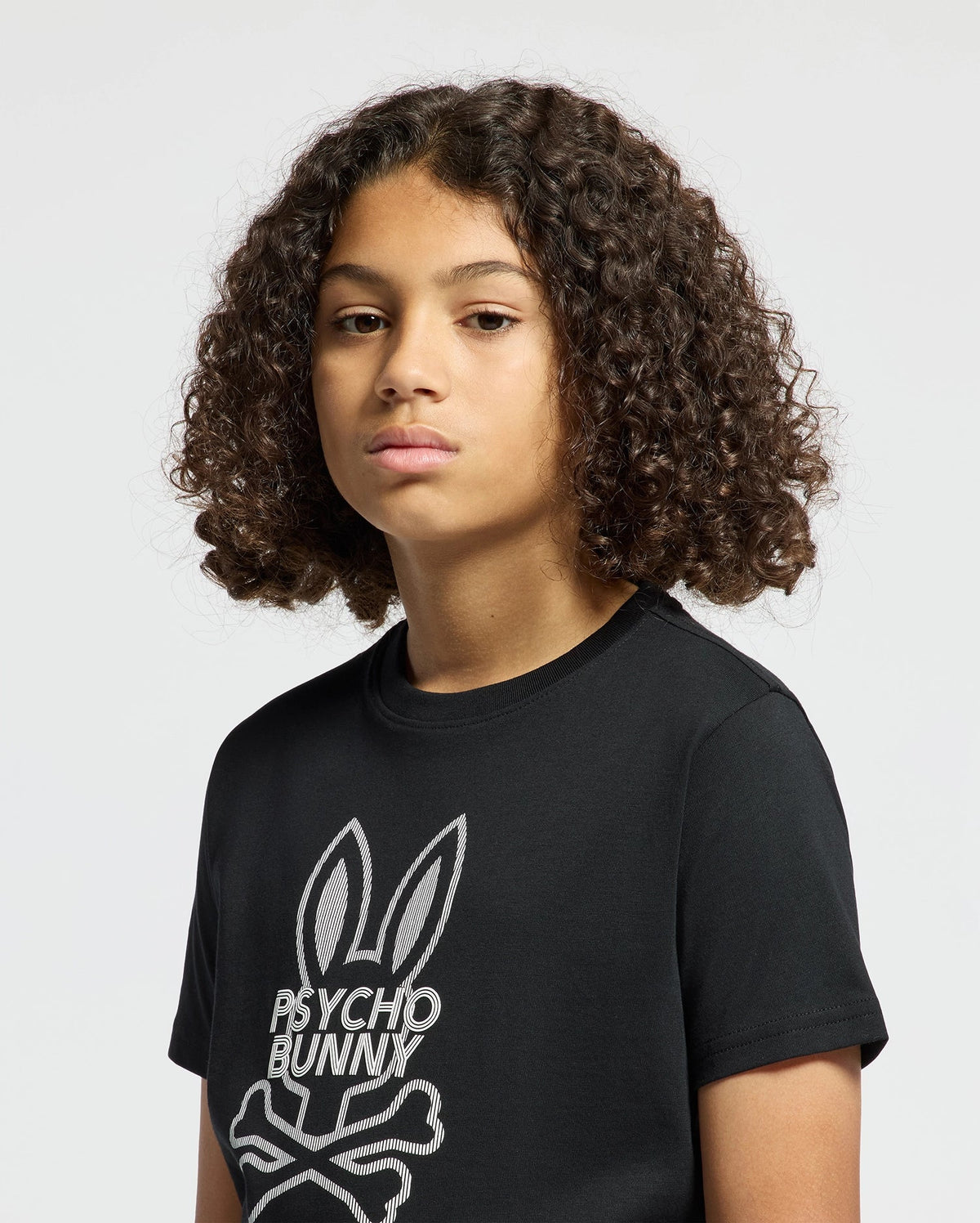 A young person with curly hair wears the trendy "KIDS DUMON GRAPHIC TEE - B0U271E200" by Psycho Bunny, showcasing a bunny and crossbones logo. The black tee, made from soft Pima cotton, enhances the calm gaze they direct at the camera against a simple backdrop.
