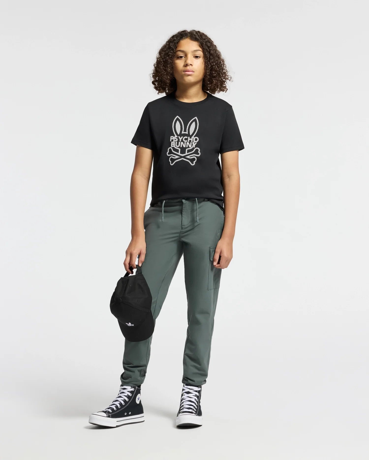 A young individual with curly hair confidently displays their trendy Psycho Bunny KIDS DUMON GRAPHIC TEE. They pair the tee with green pants and black high-top sneakers, while holding a black cap against a plain light background.