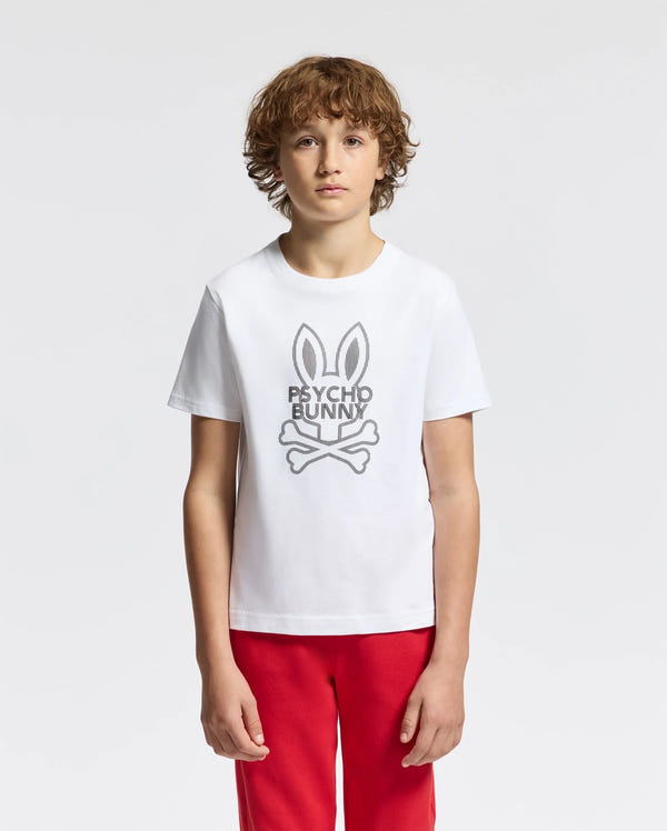 A young person with curly hair wears the KIDS DUMON GRAPHIC TEE - B0U271E200, made of Pima cotton, featuring a stylized bunny logo and "Psycho Bunny" text. They pair it with red pants against a light gray backdrop.