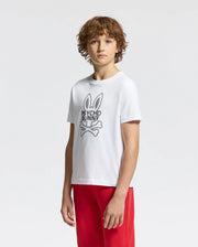 A young person with curly brown hair poses against a white backdrop, wearing the stylish KIDS DUMON GRAPHIC TEE - B0U271E200 by Psycho Bunny, made from soft Pima cotton with a bunny logo and text, paired with red pants.
