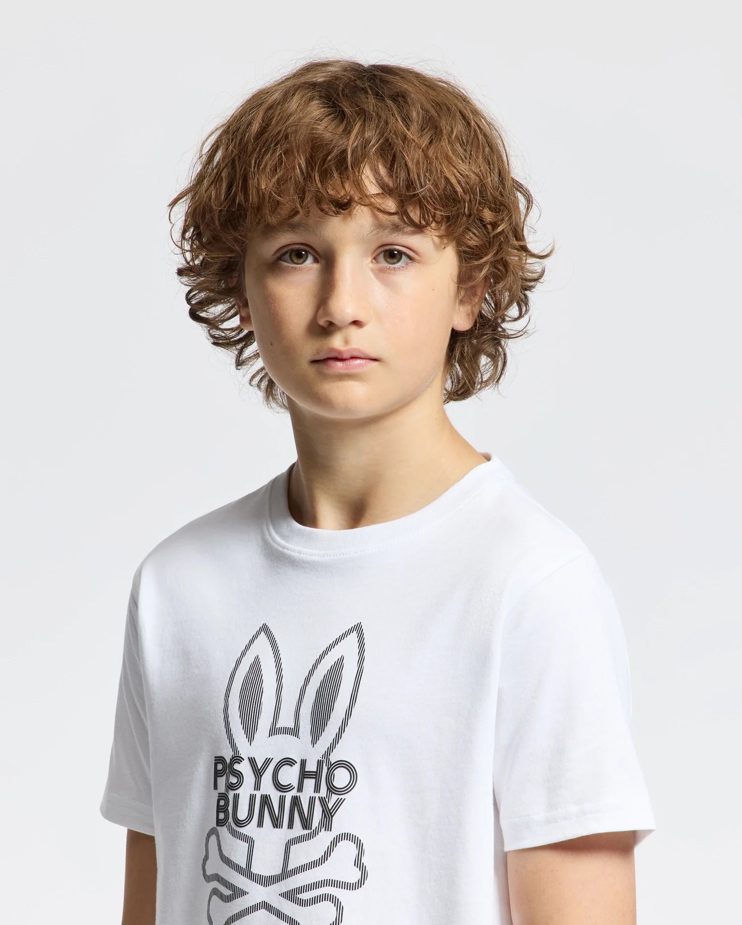 A boy with curly brown hair wears a stylish Kids Dumon Graphic Tee (B0U271E200) made of soft Pima cotton, featuring a bunny and "Psycho Bunny" text. He stands against a plain white background, looking directly at the camera.