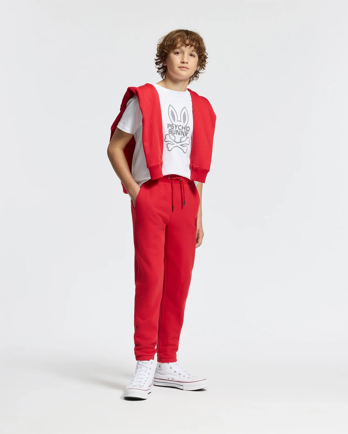 A young person with curly hair sports the stylish Kids Dumon Graphic Tee by Psycho Bunny, paired with red sweatpants and white sneakers. They drape a red sweatshirt over their shoulders, all set against a plain white background.