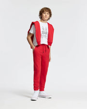 A young person with curly hair sports the stylish Kids Dumon Graphic Tee by Psycho Bunny, paired with red sweatpants and white sneakers. They drape a red sweatshirt over their shoulders, all set against a plain white background.