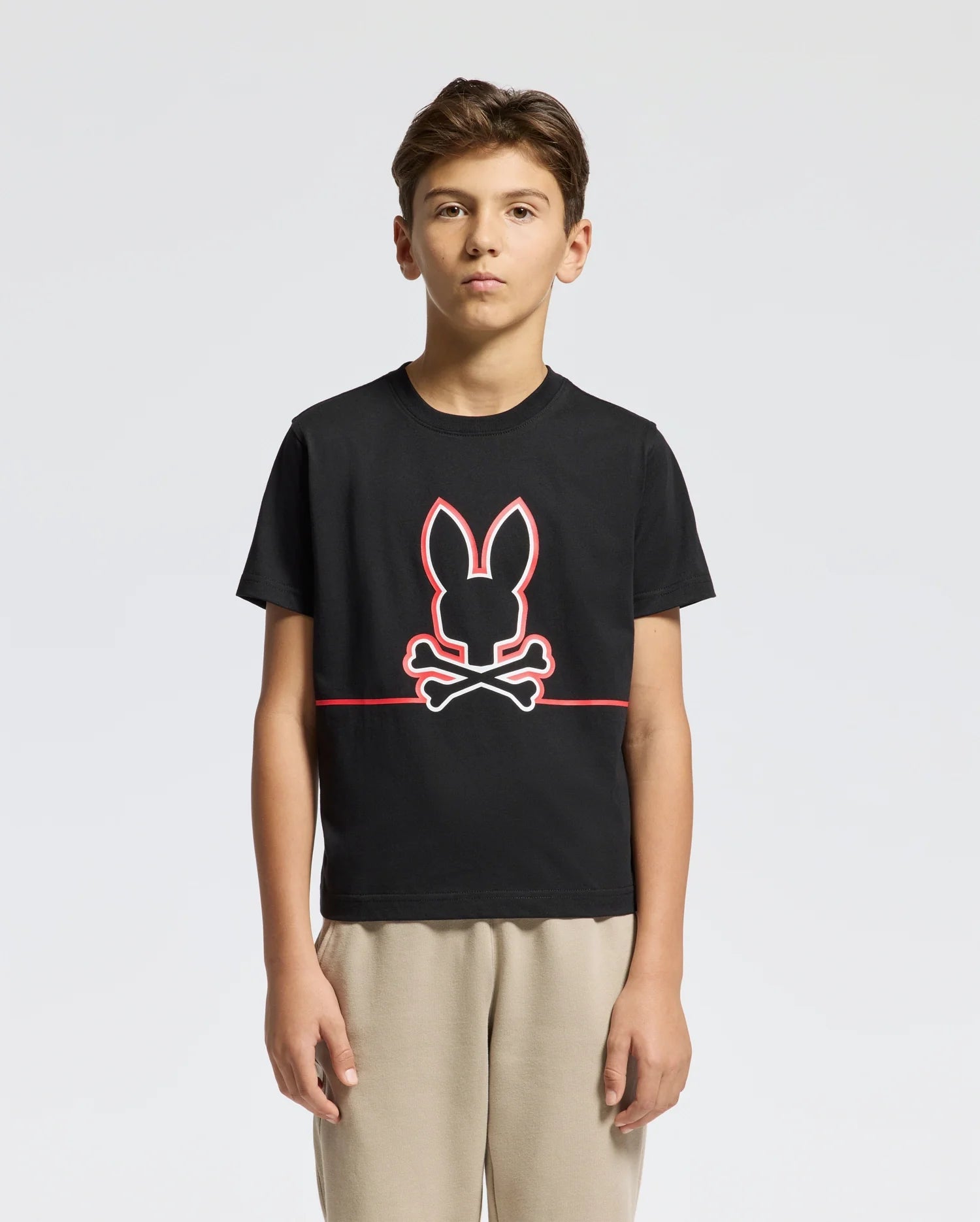 A young person stands against a plain background, wearing the black KIDS DACANO FRONT GRAPHIC TEE by Psycho Bunny, featuring a rabbit's head with crossed bones in red. Paired with regular-fit beige pants, they exude effortless style and ease.