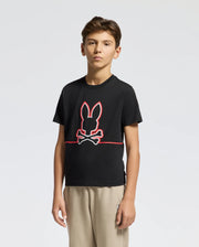 A young boy models the KIDS DACANO FRONT GRAPHIC TEE - B0U314E200 by Psycho Bunny, showcasing a red and white rabbit skull and crossbones design. With short brown hair, beige pants, and a regular-fit black Tee, his look is effortlessly stylish against a plain light background.