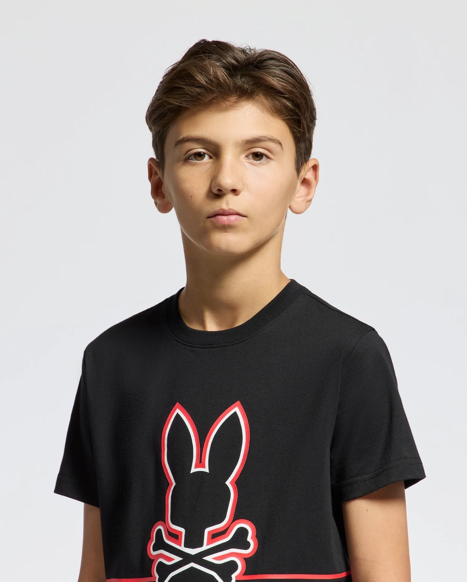 A young boy with short brown hair is wearing the Psycho Bunny KIDS DACANO FRONT GRAPHIC TEE - B0U314E200. The black regular fit tee with a bunny and crossbones design outlined in red offers both style and comfort as he poses against a plain white background.