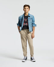 A young person wears a blue denim jacket and the Psycho Bunny KIDS DACANO FRONT GRAPHIC TEE - B0U314E200, featuring a red and white logo, paired with beige regular-fit joggers. Black high-top sneakers complete the look as they stand with hands in pockets against a plain background.