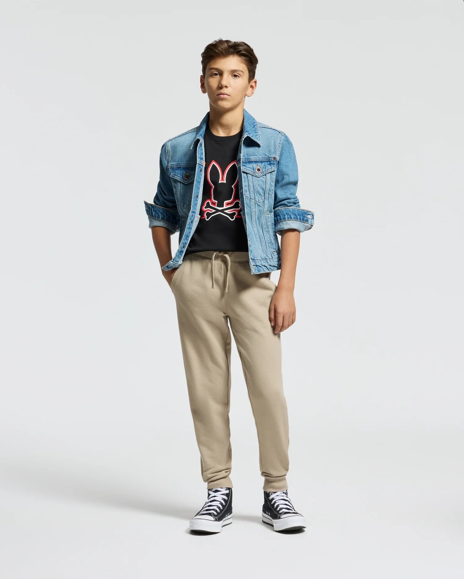 A young person wears a blue denim jacket and the Psycho Bunny KIDS DACANO FRONT GRAPHIC TEE - B0U314E200, featuring a red and white logo, paired with beige regular-fit joggers. Black high-top sneakers complete the look as they stand with hands in pockets against a plain background.