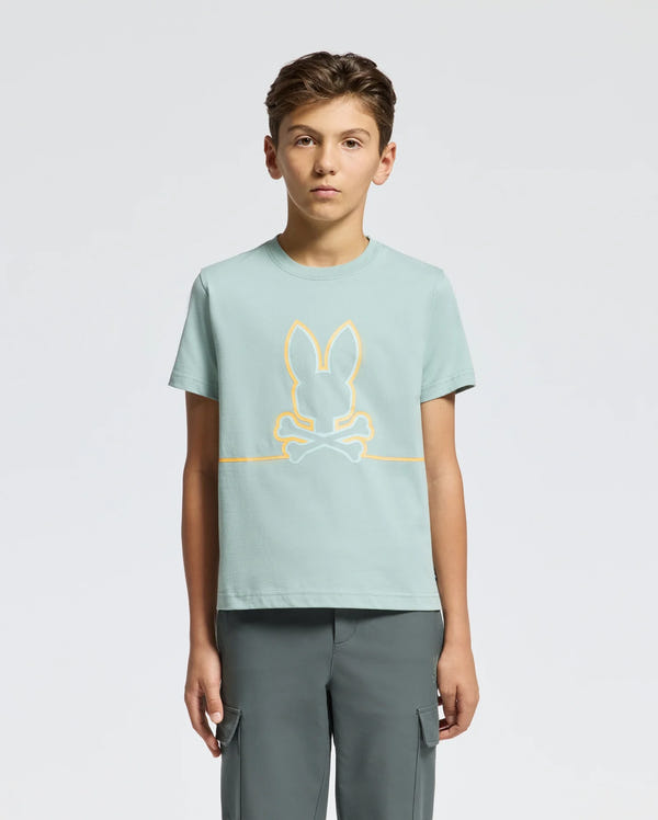 A teen in a comfort-focused, light green KIDS DACANO FRONT GRAPHIC TEE - B0U314E200 by Psycho Bunny featuring a bunny and crossbones design pairs it with dark green pants. Their short, light brown hair contrasts nicely against the plain white background.