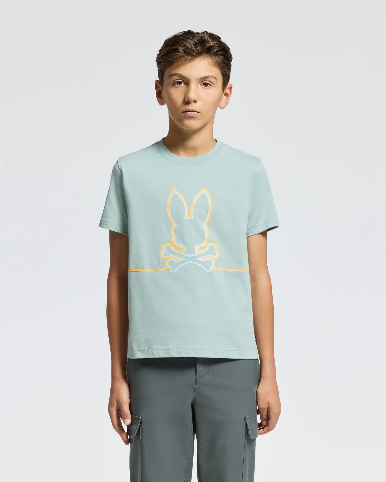 A teen in a comfort-focused, light green KIDS DACANO FRONT GRAPHIC TEE - B0U314E200 by Psycho Bunny featuring a bunny and crossbones design pairs it with dark green pants. Their short, light brown hair contrasts nicely against the plain white background.
