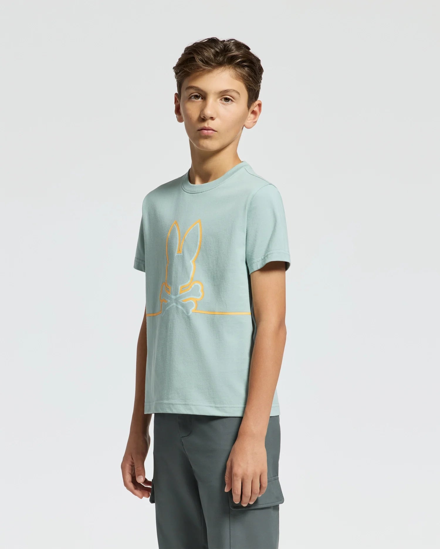 A teen boy stands against a plain background, showcasing the Psycho Bunny KIDS DACANO FRONT GRAPHIC TEE. The light green tee, designed for comfort with an orange rabbit and bone outline, features a regular fit that emphasizes casual style. His hands are by his sides as he looks forward.