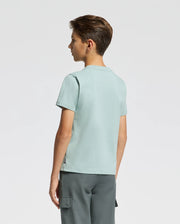 A young person with short hair stands in profile, wearing a stylishly comfortable Psycho Bunny KIDS DACANO FRONT GRAPHIC TEE in light blue paired with dark gray cargo pants against a plain white background.