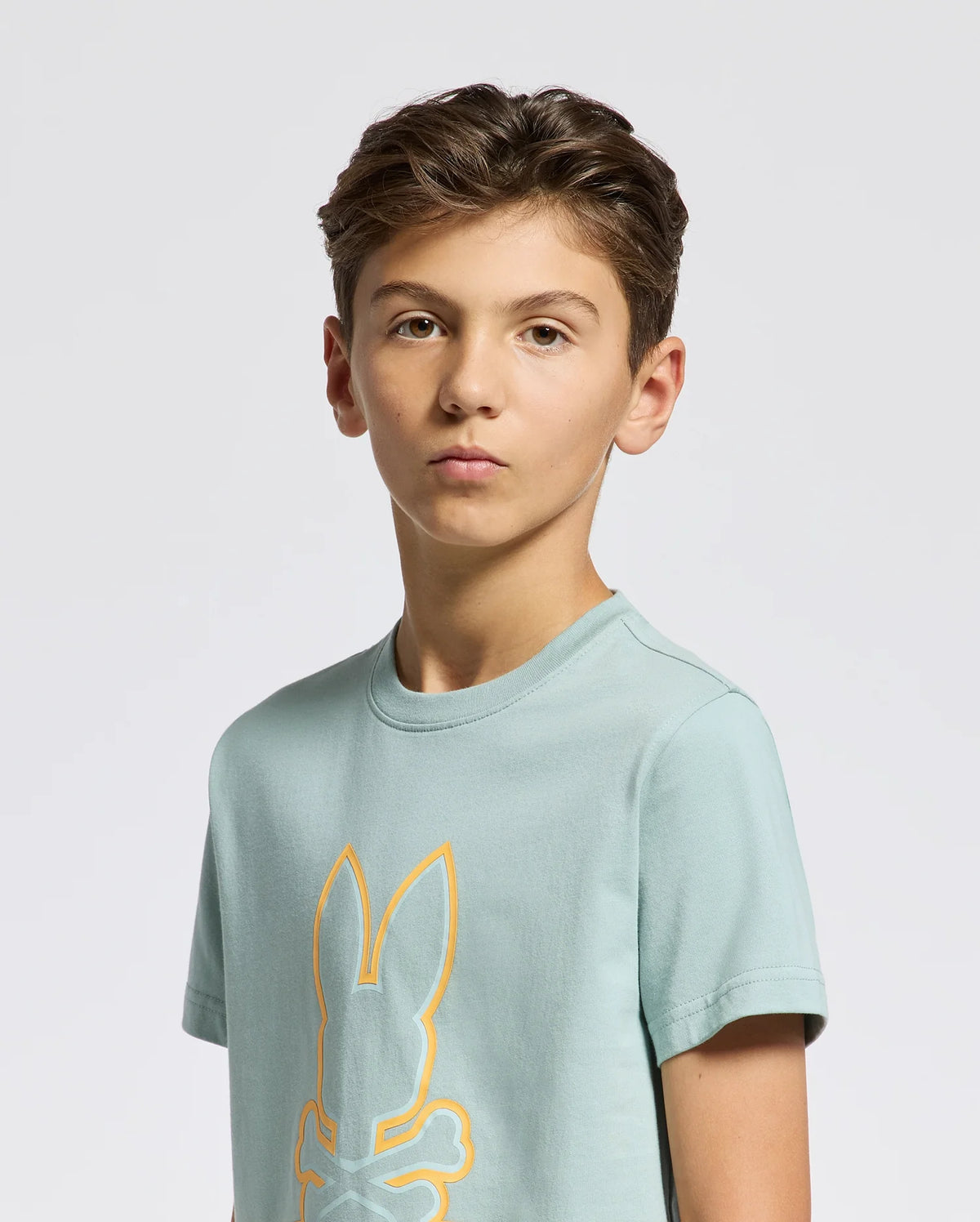 A young person poses against a white backdrop in the Psycho Bunny KIDS DACANO FRONT GRAPHIC TEE - B0U314E200, a comfort-forward light blue shirt. Featuring a vibrant yellow bunny and crossbones design, this regular fit tee offers style and ease for any casual outing.