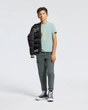 A teen sports a light blue KIDS DACANO FRONT GRAPHIC TEE from Psycho Bunny, effortlessly stylish with olive cargo pants, black high-tops, and a black bomber jacket draped over his shoulder against a plain gray backdrop.