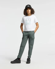 A person with curly hair is wearing a black cap, the stylish Psycho Bunny KIDS CORTES GRAPHIC TEE - B0U328E200 made from 100% cotton, and green cargo pants. They stand with hands in pockets, sporting black sneakers against a plain white background.