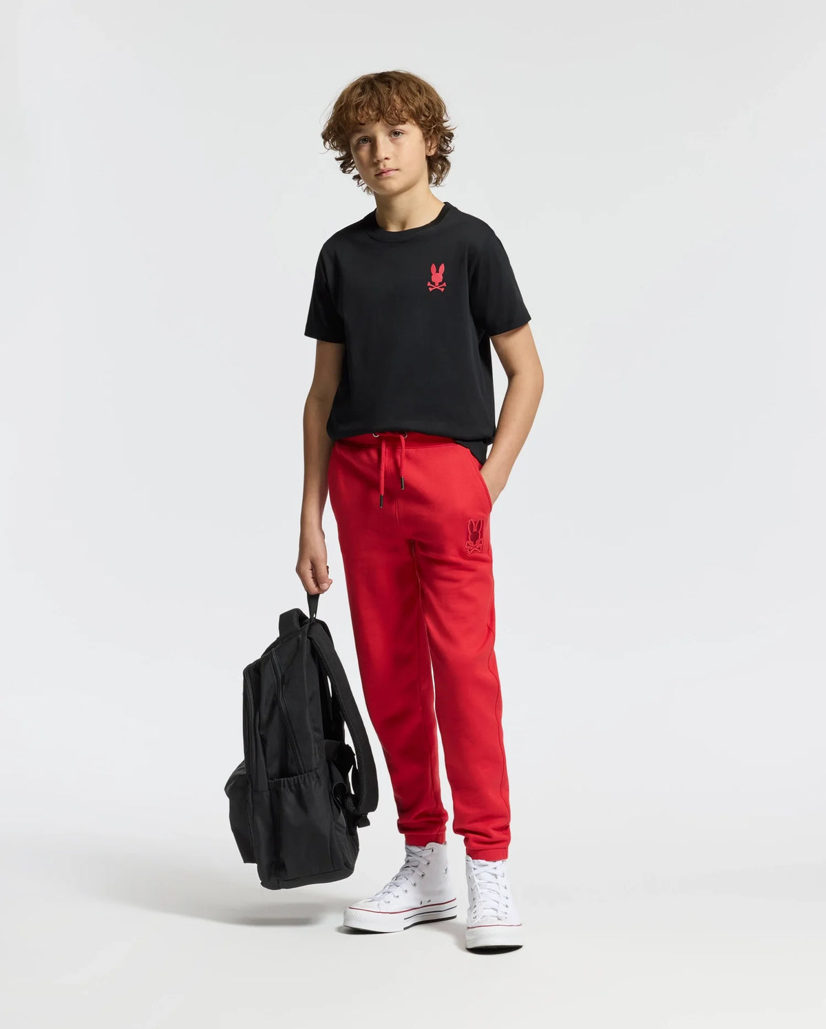 A young person with curly hair wears a stylish Psycho Bunny black graphic tee featuring a red logo from the KIDS RIO collection, paired with red sweatpants and white high-top sneakers. They are holding a black backpack and standing against a plain white background.