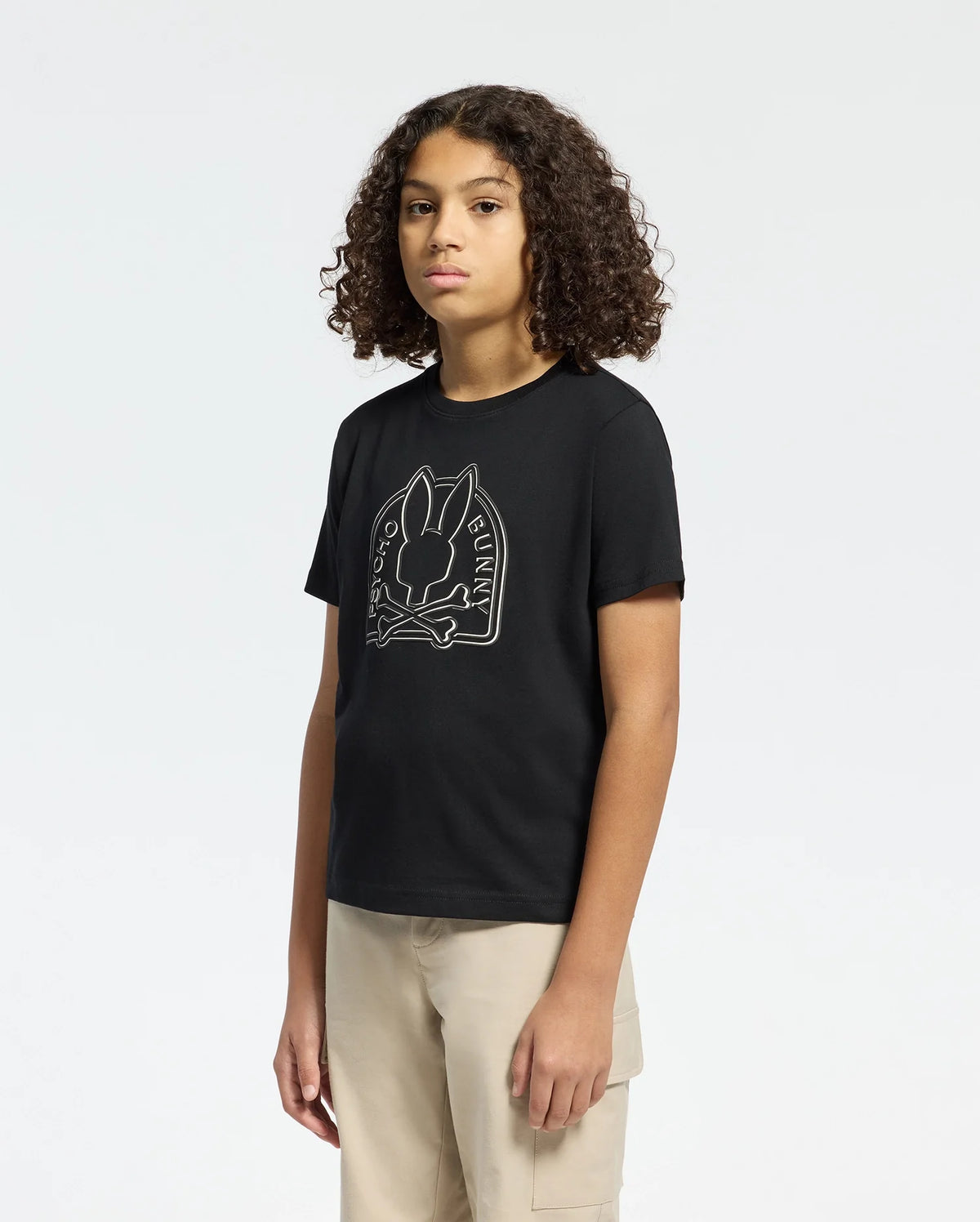 A person with curly hair in the KIDS MELBOURNE GRAPHIC TEE - B0U410F200 by Psycho Bunny, featuring a bunny graphic and text, pairs it with beige pants against a plain white background.
