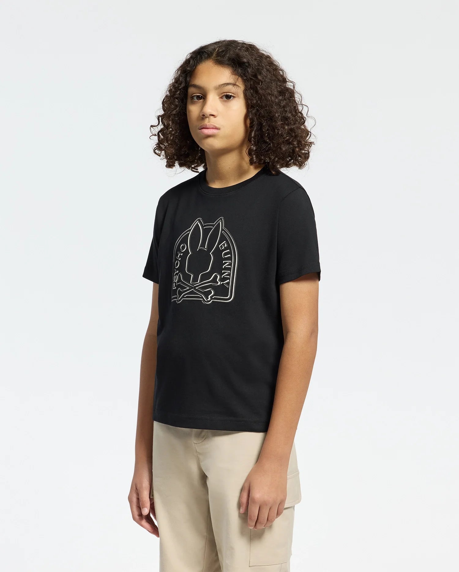 A person with curly hair in the KIDS MELBOURNE GRAPHIC TEE - B0U410F200 by Psycho Bunny, featuring a bunny graphic and text, pairs it with beige pants against a plain white background.