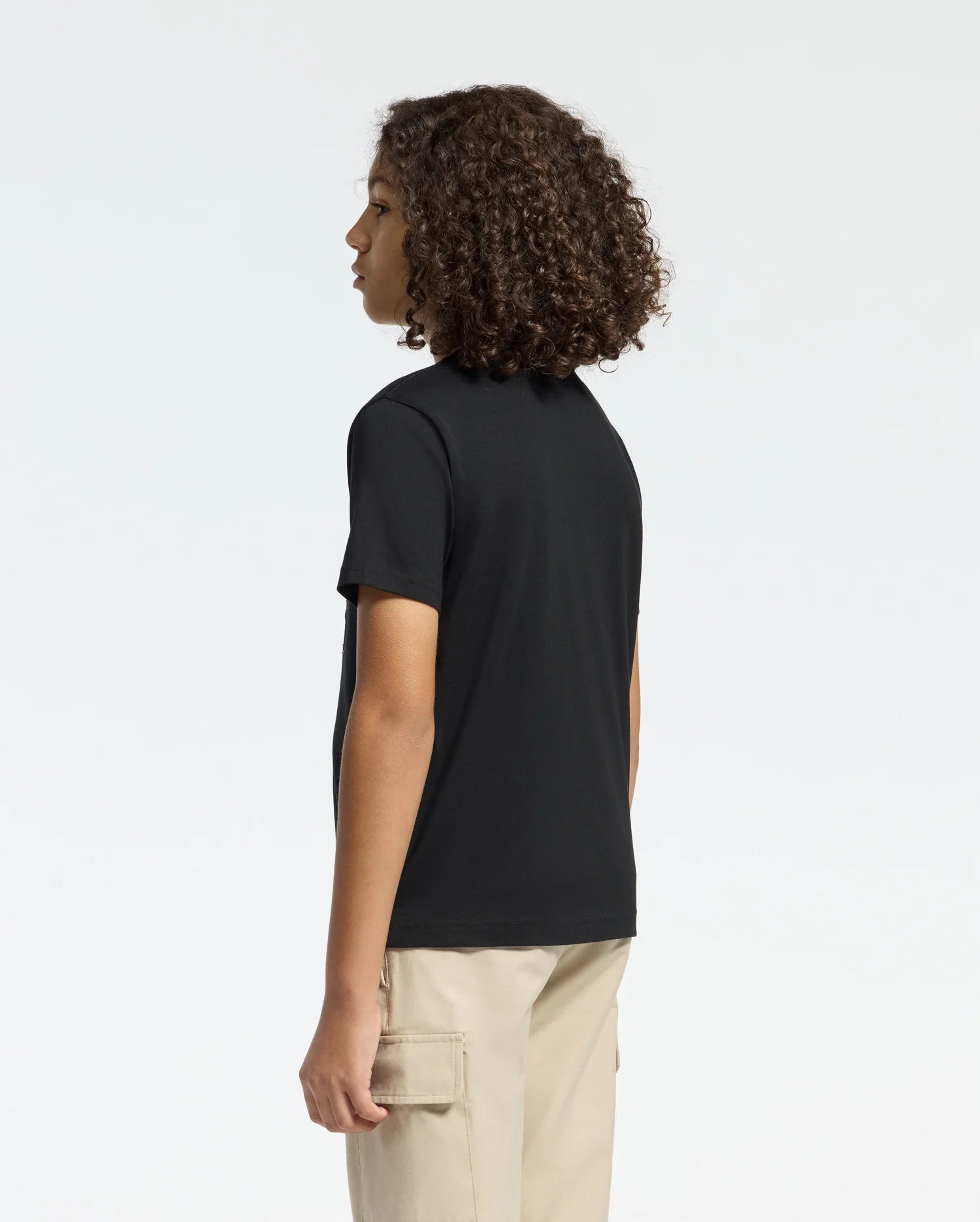 A person with curly hair stands sideways, facing left, wearing a black KIDS MELBOURNE GRAPHIC TEE by Psycho Bunny and beige cargo pants. The plain white background enhances the striking simplicity of their Pima cotton outfit.