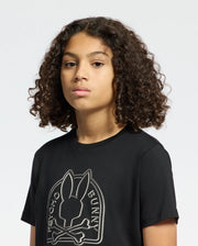 A person with curly hair sports a Kids Melbourne Graphic Tee by Psycho Bunny, made from soft Pima cotton. It showcases a striking bunny graphic—a rabbit skull and crossbones with the text "Bunny Psycho"—on a plain white background.