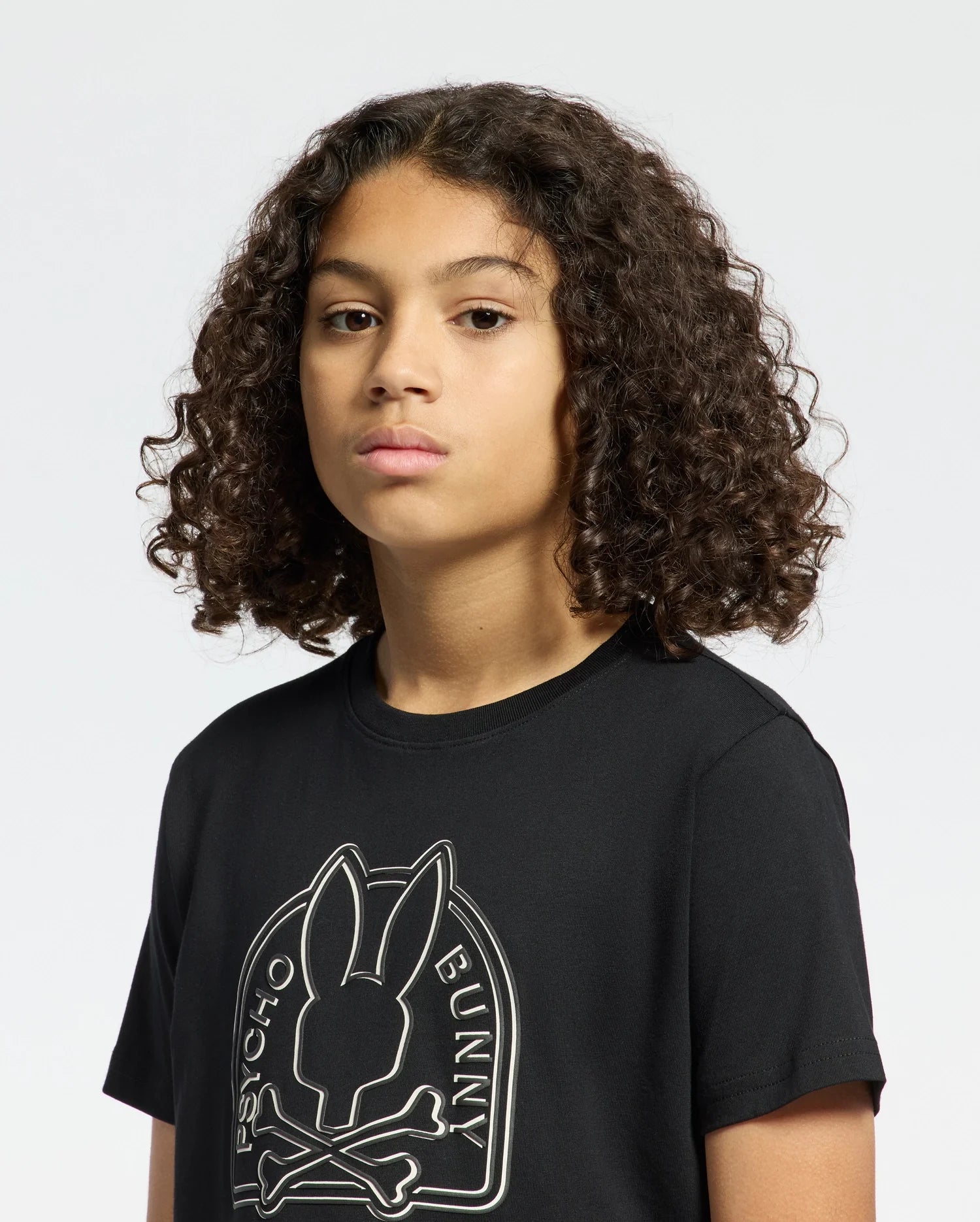 A person with curly hair sports a Kids Melbourne Graphic Tee by Psycho Bunny, made from soft Pima cotton. It showcases a striking bunny graphic—a rabbit skull and crossbones with the text "Bunny Psycho"—on a plain white background.