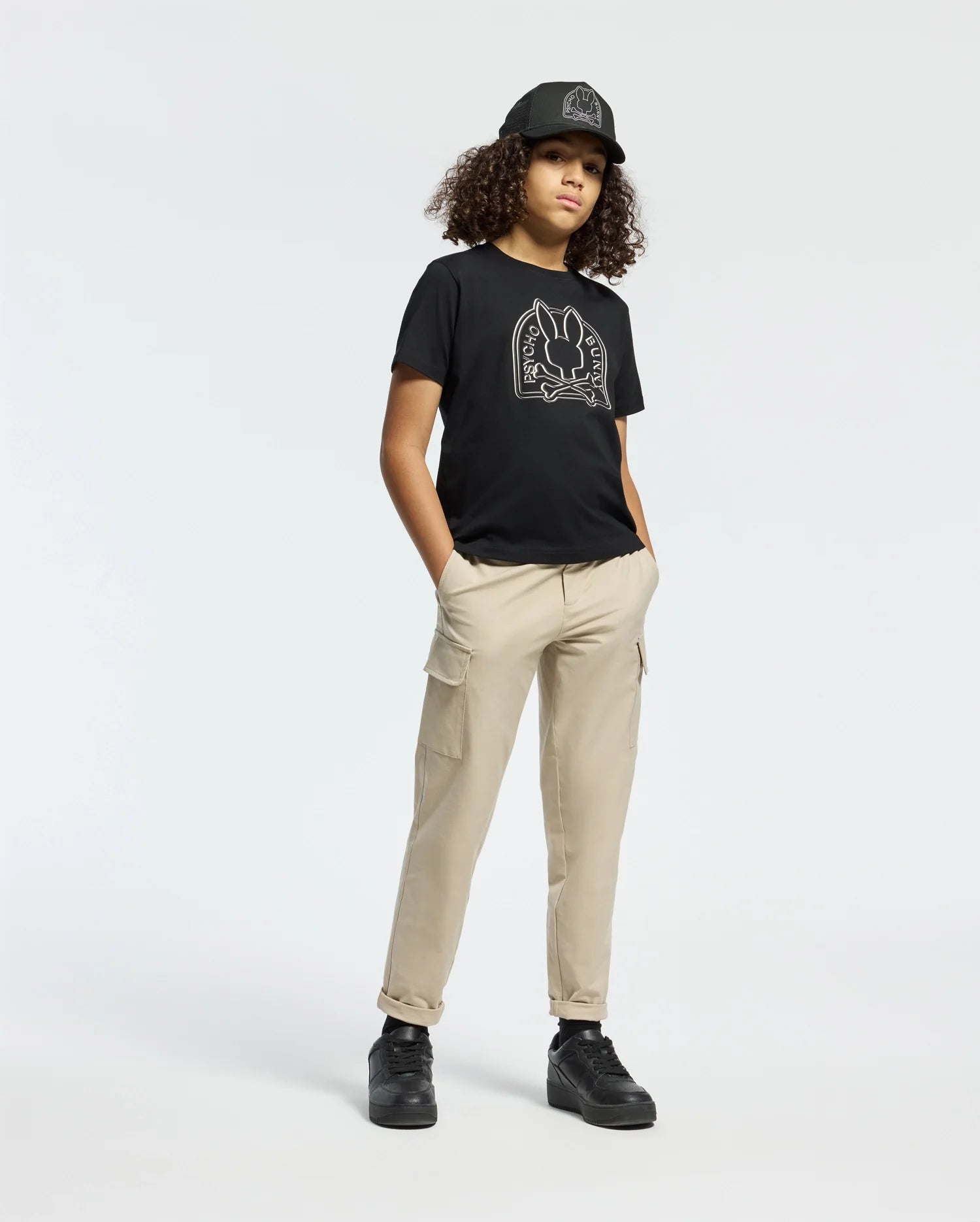 A person with curly hair is wearing a black cap, Psycho Bunny's Kids Melbourne Graphic Tee (B0U410F200) with a bunny graphic, beige cargo pants, and black sneakers while standing against a plain white background with hands in their pockets.