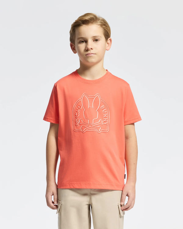 A young boy wears a vibrant orange KIDS MELBOURNE GRAPHIC TEE (B0U410F200) made of soft Peruvian Pima cotton, featuring a striking bunny graphic with crossed bones and "Psycho Bunny" branding. He pairs it with beige pants, confidently standing against a plain white background.