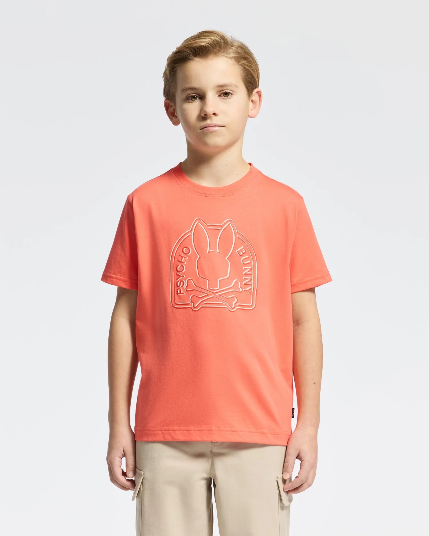 A young boy wears a vibrant orange KIDS MELBOURNE GRAPHIC TEE (B0U410F200) made of soft Peruvian Pima cotton, featuring a striking bunny graphic with crossed bones and "Psycho Bunny" branding. He pairs it with beige pants, confidently standing against a plain white background.
