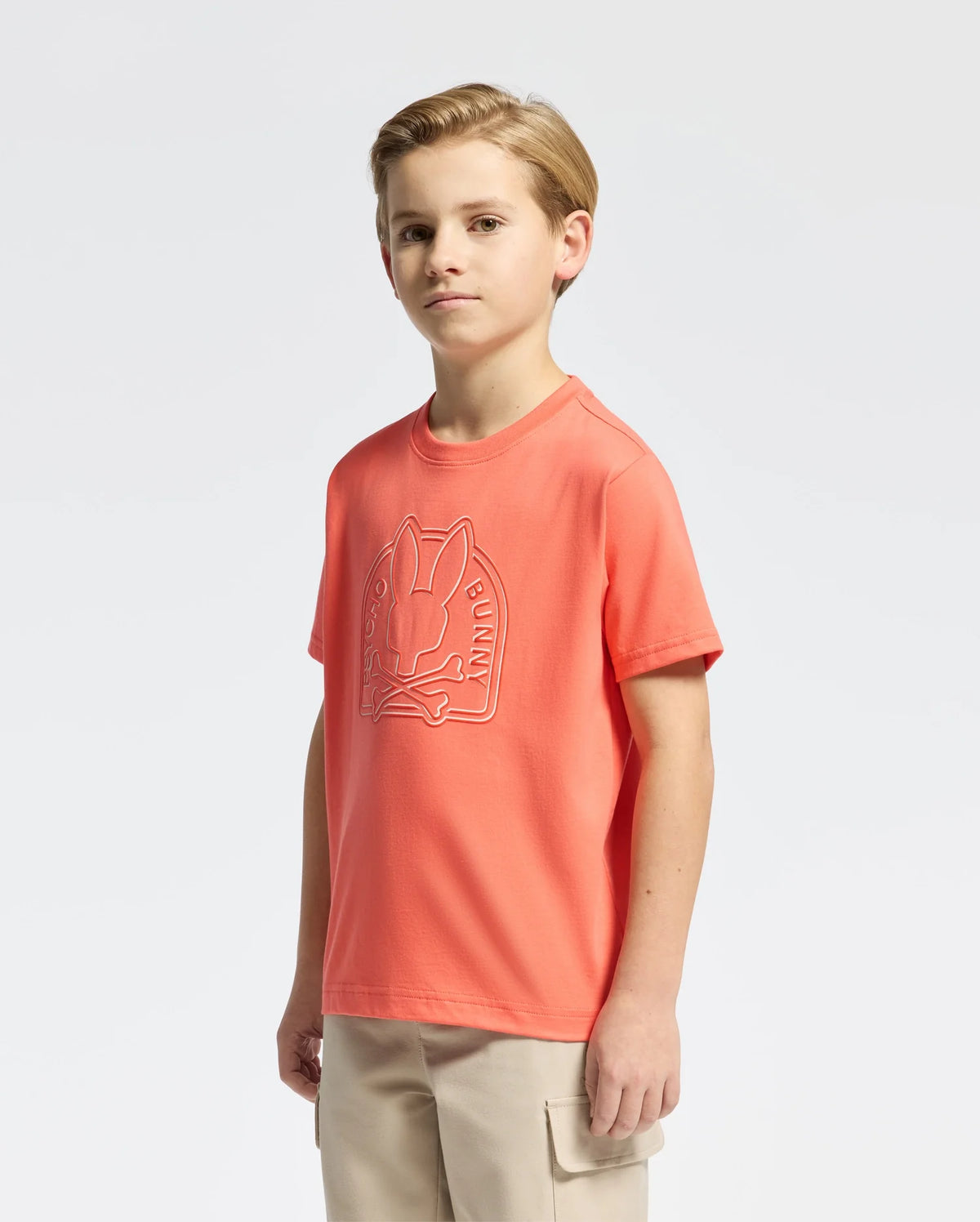 A young person faces forward wearing the Psycho Bunny KIDS MELBOURNE GRAPHIC TEE - B0U410F200, an orange tee made of soft Peruvian Pima cotton with a bunny graphic, paired with beige pants against a plain white background.