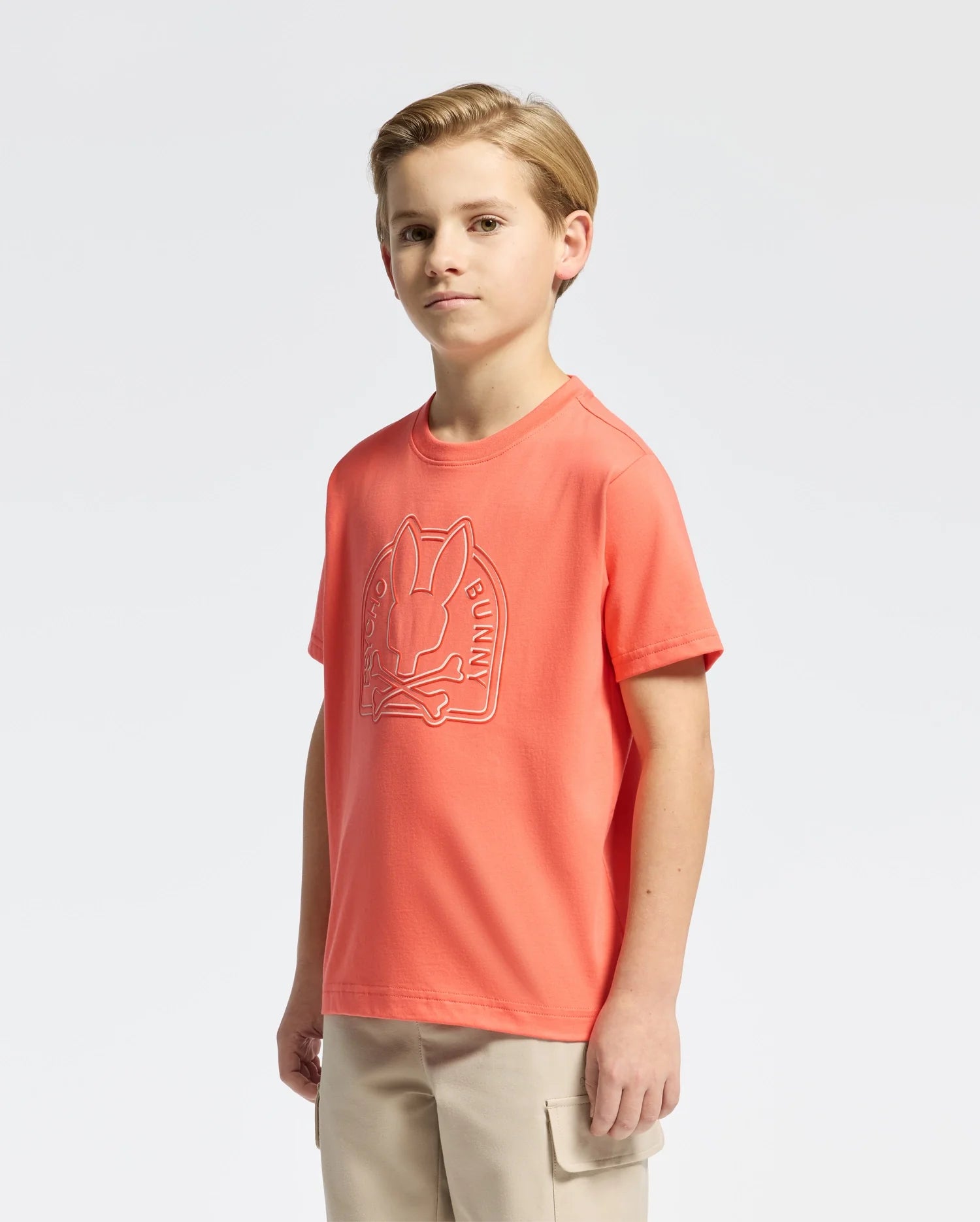 A young person faces forward wearing the Psycho Bunny KIDS MELBOURNE GRAPHIC TEE - B0U410F200, an orange tee made of soft Peruvian Pima cotton with a bunny graphic, paired with beige pants against a plain white background.