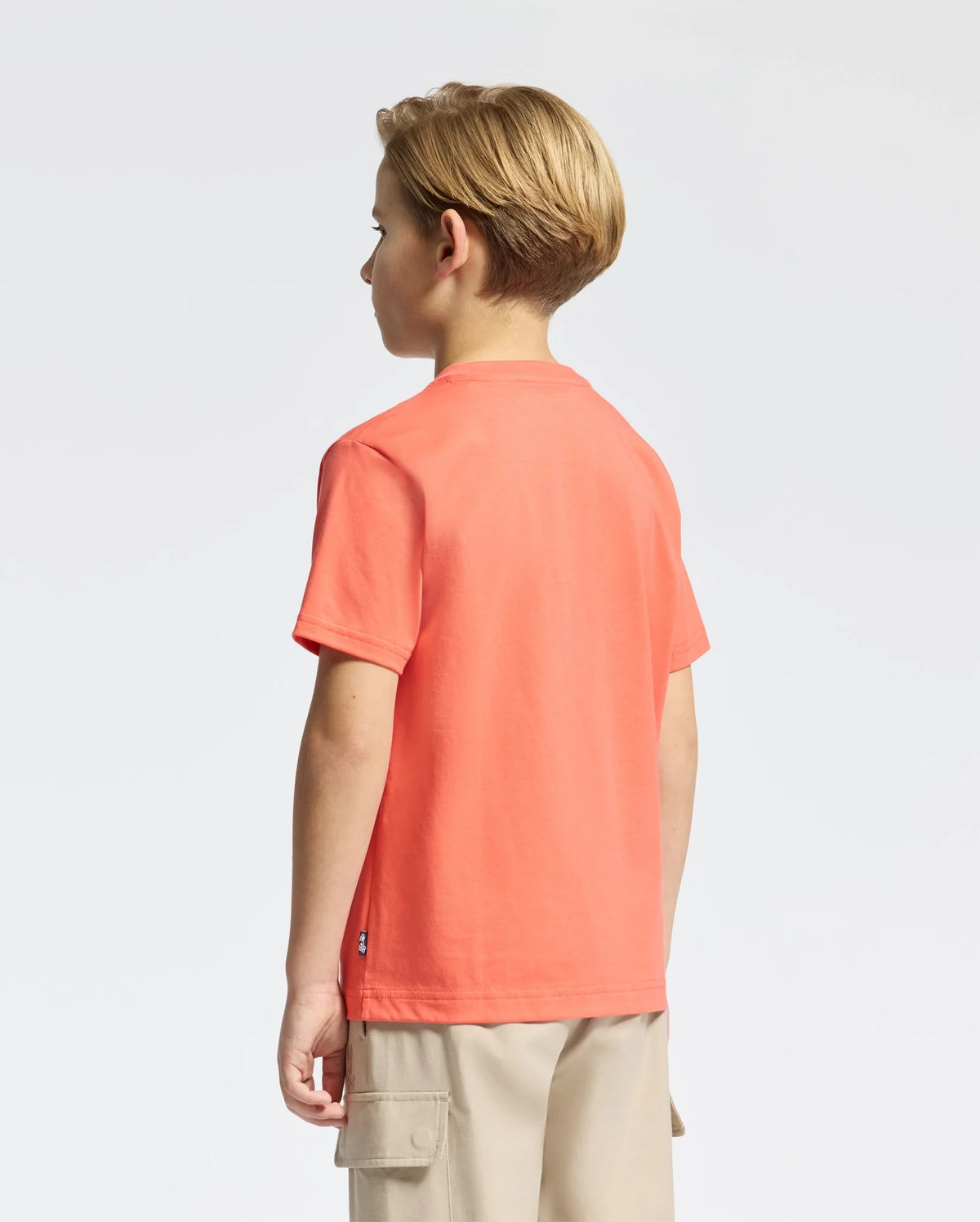 A young blonde child faces away, wearing an orange KIDS MELBOURNE GRAPHIC TEE by Psycho Bunny crafted from soft Peruvian Pima cotton, paired with beige cargo shorts, against a plain white backdrop.