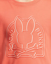 The KIDS MELBOURNE GRAPHIC TEE - B0U410F200 by Psycho Bunny features a bunny with elongated ears over crossbones, crafted from Peruvian Pima cotton. The words "Psycho Bunny" arch above this coral-colored t-shirt design.