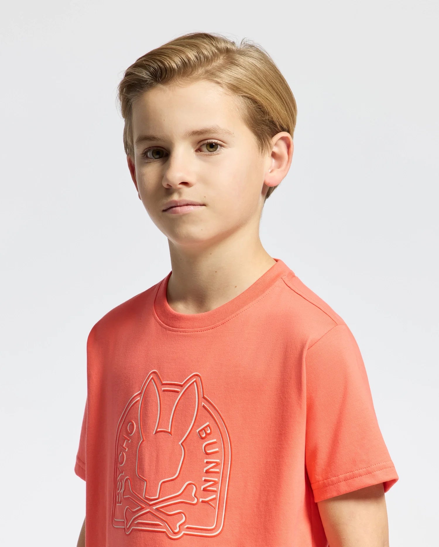 A young boy with short, light brown hair is wearing the Psycho Bunny KIDS MELBOURNE GRAPHIC TEE (B0U410F200) in vibrant orange, featuring a bunny graphic with crossbones. Made from soft Peruvian Pima cotton, he stands against a light background, gazing slightly to the right.