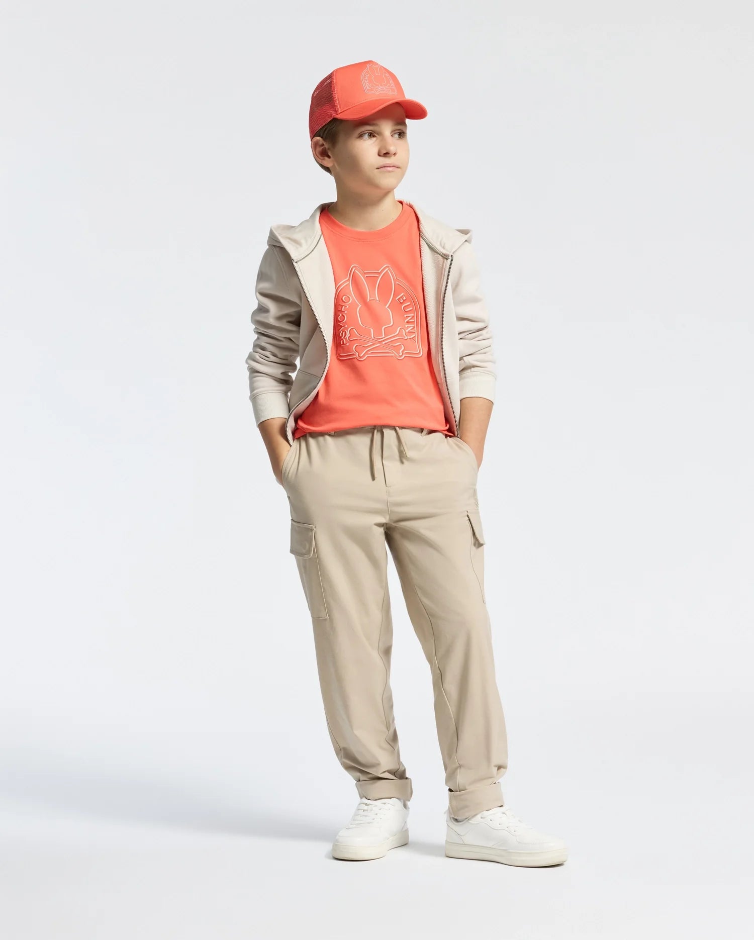 A young boy sports a coral cap, Psycho Bunny KIDS MELBOURNE GRAPHIC TEE featuring a bunny design, beige joggers, and a light beige zip-up hoodie. He stands with hands in pockets, gazing to the side against a plain white backdrop.