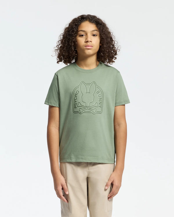 A person with curly hair wears a Psycho Bunny sage green KIDS MELBOURNE GRAPHIC TEE - B0U410F200, showcasing a large bunny and crossbones water-based print on the front. They pair it with beige pants against a plain white background.