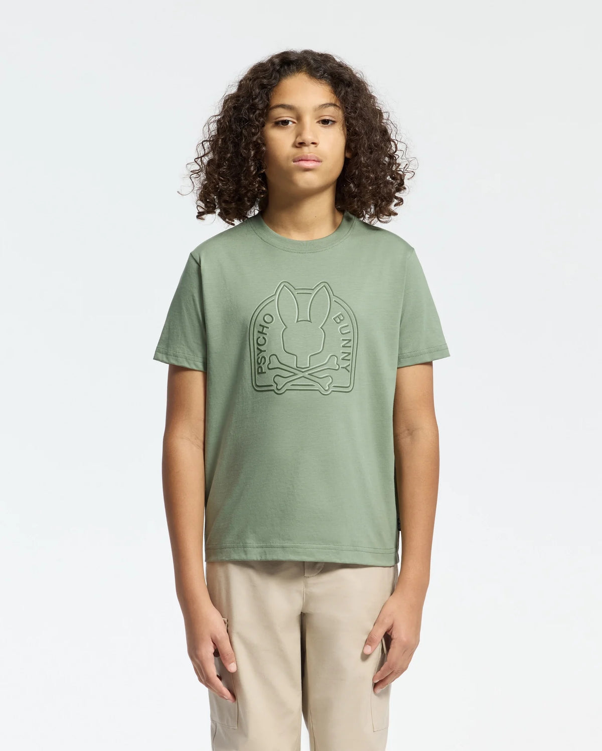 A person with curly hair wears a Psycho Bunny sage green KIDS MELBOURNE GRAPHIC TEE - B0U410F200, showcasing a large bunny and crossbones water-based print on the front. They pair it with beige pants against a plain white background.