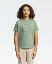 A person with curly hair wears a Psycho Bunny sage green KIDS MELBOURNE GRAPHIC TEE - B0U410F200, showcasing a large bunny and crossbones water-based print on the front. They pair it with beige pants against a plain white background.
