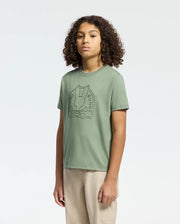 A curly-haired individual wears Psycho Bunny's KIDS MELBOURNE GRAPHIC TEE in sage green, made of soft Pima cotton with a bunny design. They pair it with beige pants against a white background.