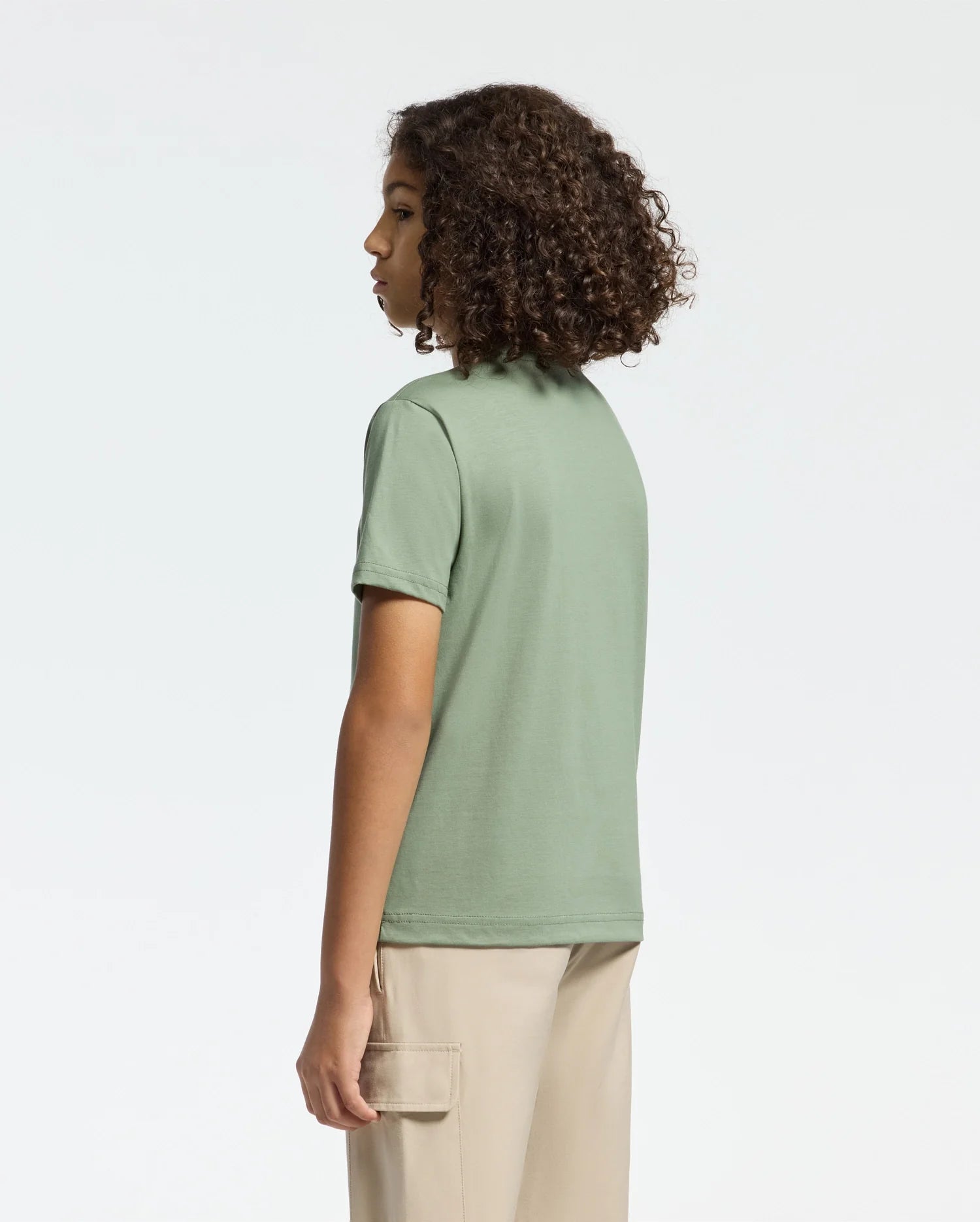 A curly-haired person, wearing the Psycho Bunny KIDS MELBOURNE GRAPHIC TEE in green Pima cotton with a bunny print, poses sideways against a plain backdrop. Their beige pants offer an understated complement to the look.