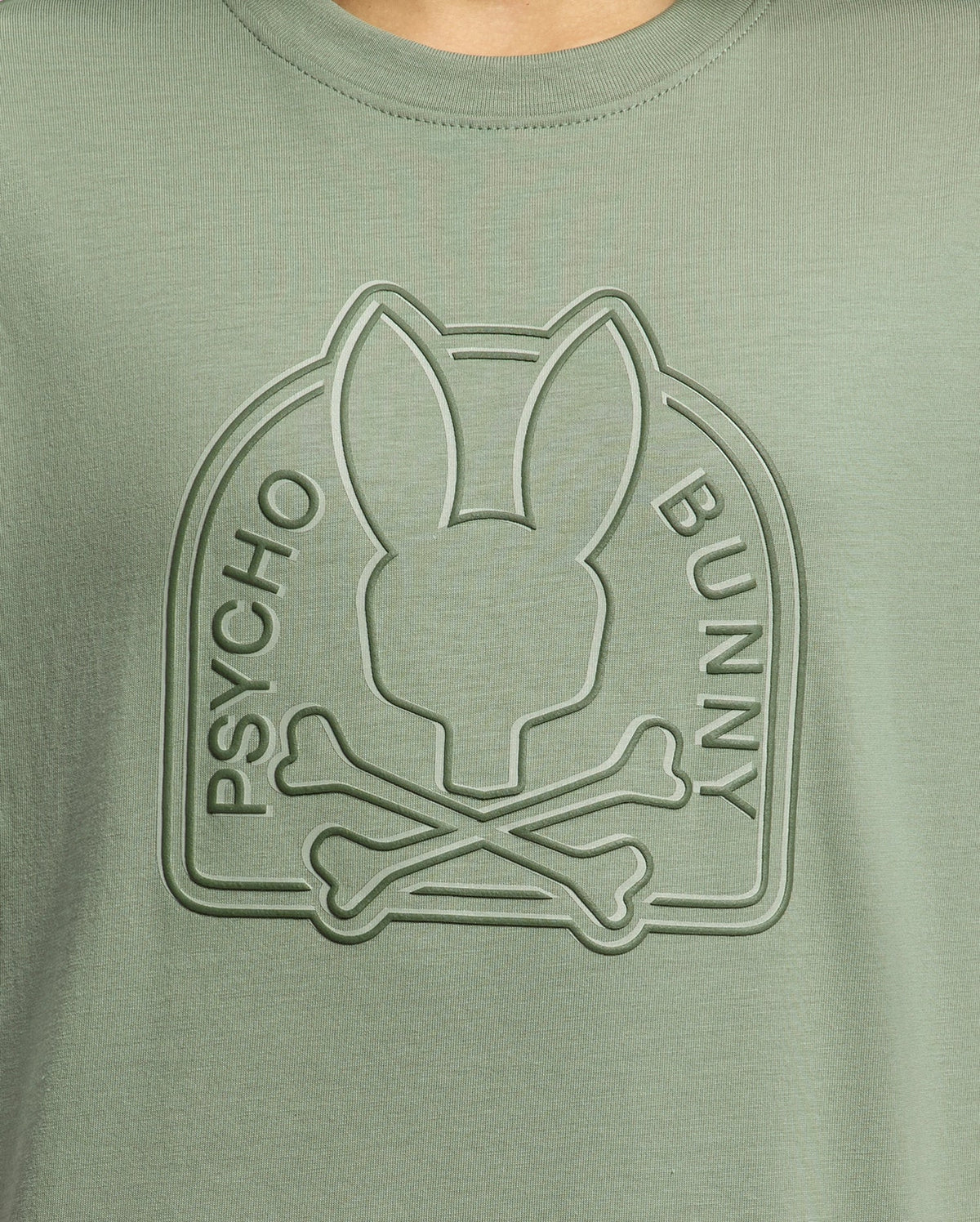Close-up of the Psycho Bunny Kids Melbourne Graphic Tee in green Pima cotton, featuring a raised water-based print with a stylized bunny head atop crossed bones within an arch, with "Psycho Bunny" embossed on the arch's sides.