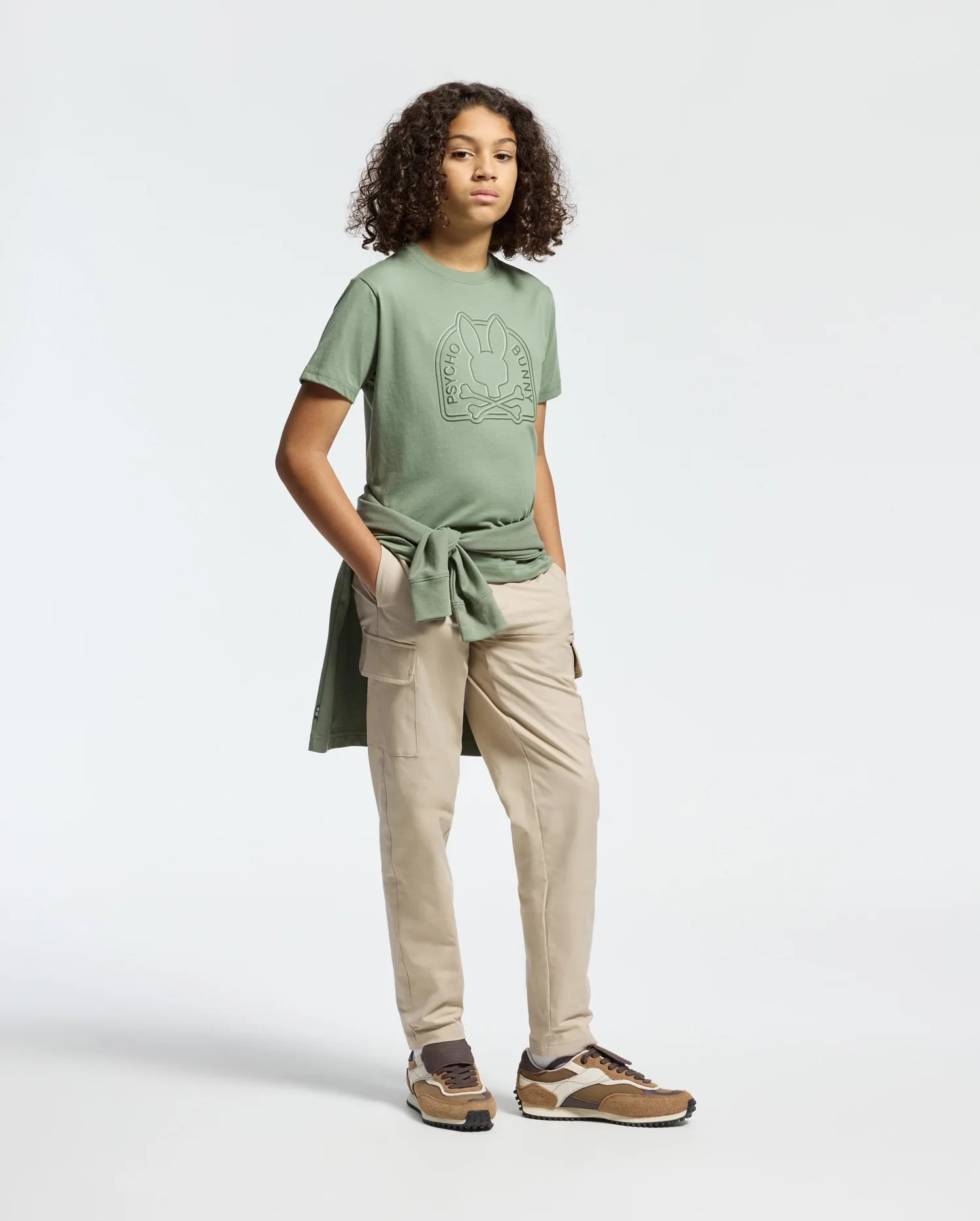 A person with curly hair wears Psycho Bunny's KIDS MELBOURNE GRAPHIC TEE in green with a water-based bunny print. They're styled in light beige cargo pants, brown sneakers, and have a matching green Pima cotton sweatshirt around their waist, against a plain light background.