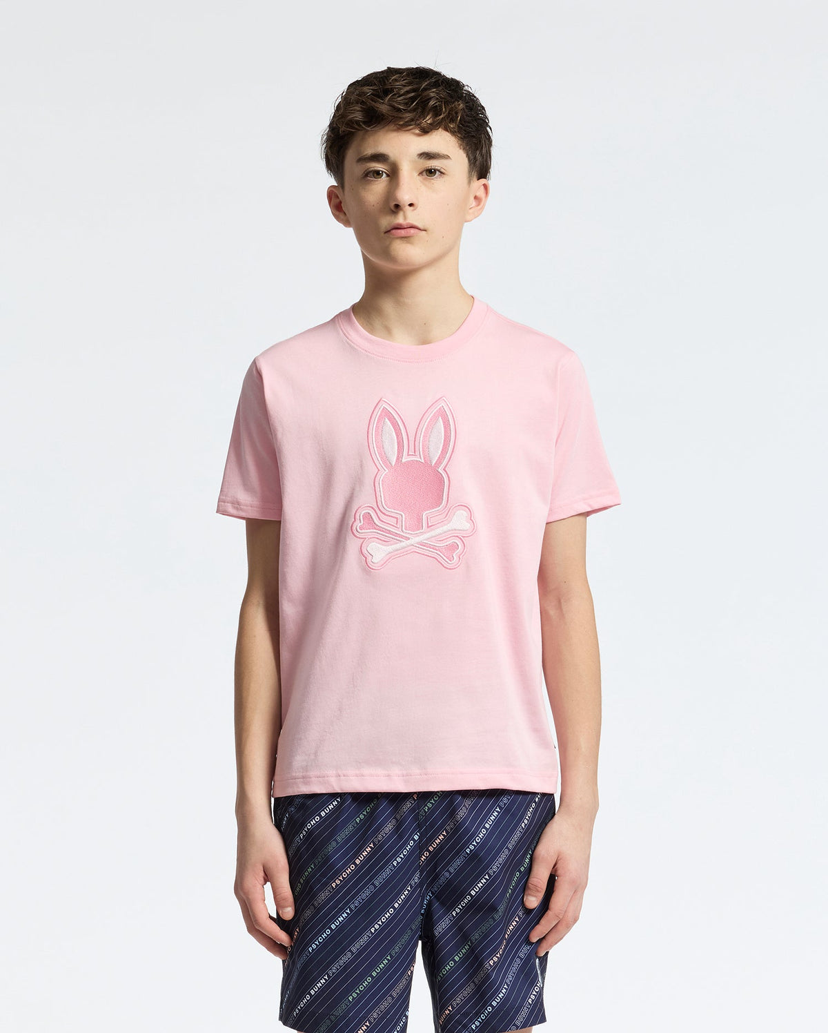 A teen boy wears the Psycho Bunny KIDS NEW CASTLE EMBROIDERED GRAPHIC TEE (B0U415F200) in pink, made from soft Pima cotton with a bunny and crossbones design. He pairs it effortlessly with navy patterned shorts, standing against a plain white background.