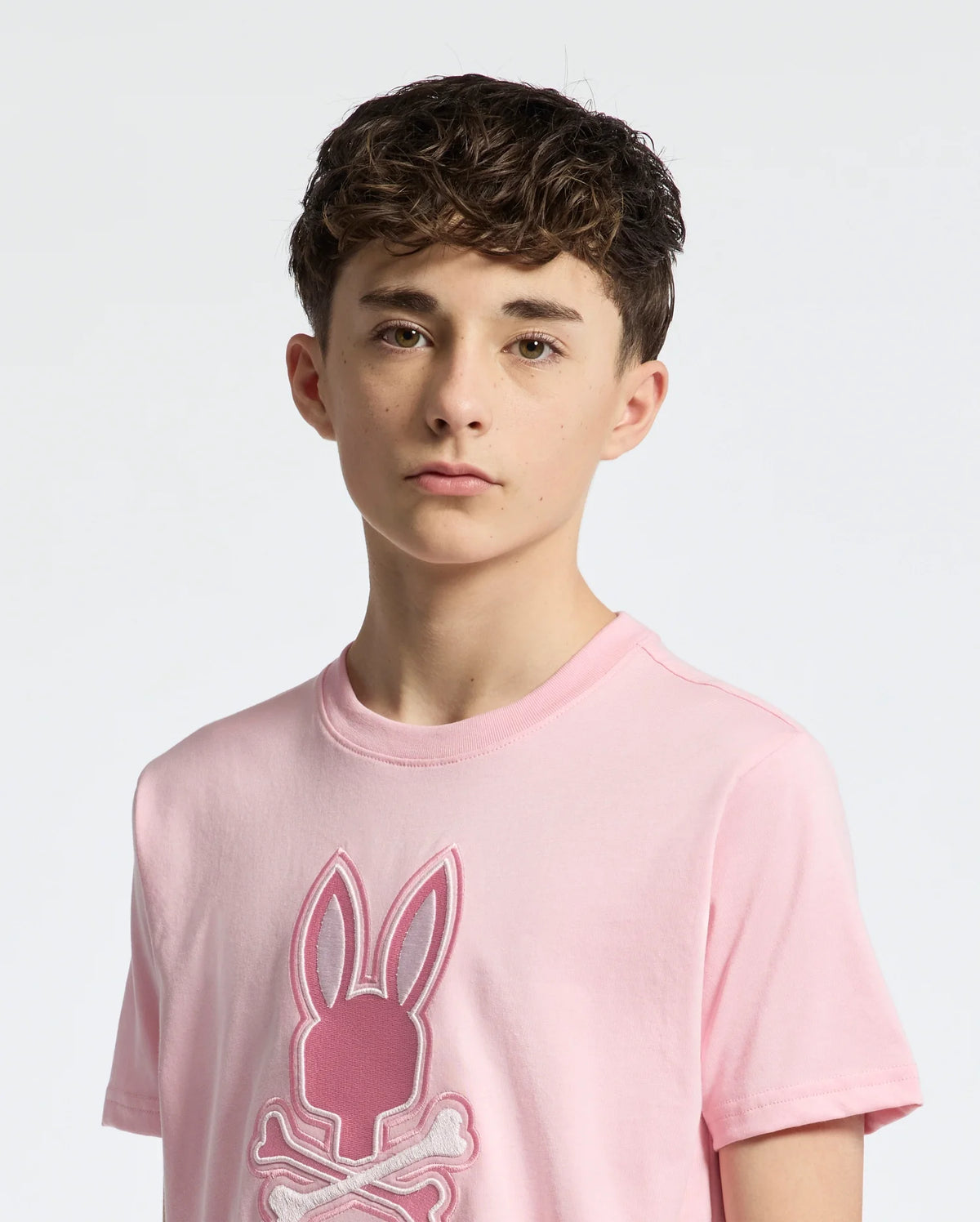 A teen with short, curly brown hair wears Psycho Bunny's KIDS NEW CASTLE EMBROIDERED GRAPHIC TEE (B0U415F200), a pink Pima cotton tee featuring an embroidered bunny and crossbones. He stands against a white background, looking directly at the camera with a neutral expression.
