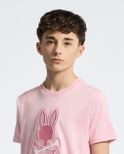 A teen with short, curly brown hair wears Psycho Bunny's KIDS NEW CASTLE EMBROIDERED GRAPHIC TEE (B0U415F200), a pink Pima cotton tee featuring an embroidered bunny and crossbones. He stands against a white background, looking directly at the camera with a neutral expression.