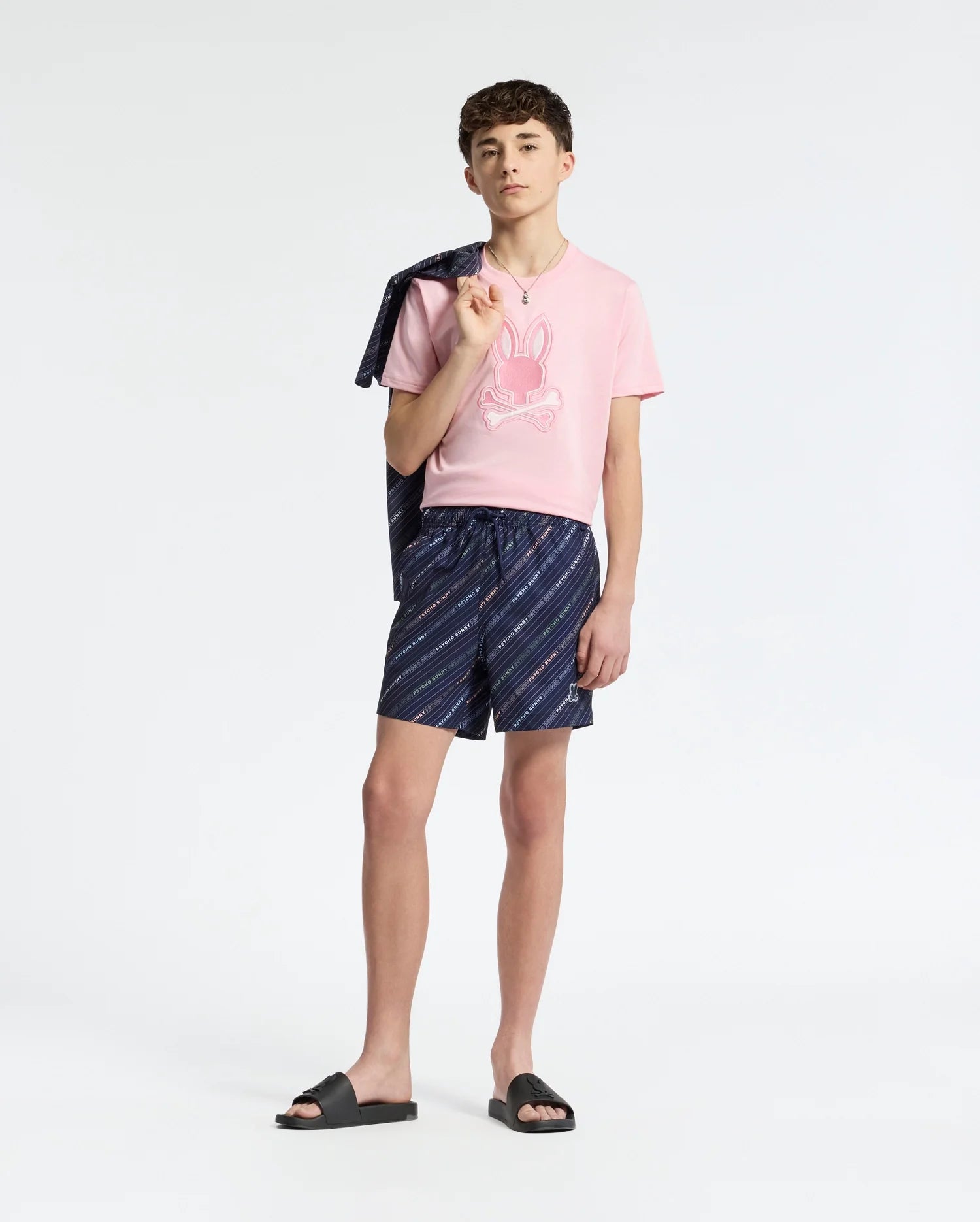 A teen boy stands against a white background wearing a Psycho Bunny pink tee, KIDS NEW CASTLE EMBROIDERED GRAPHIC TEE - B0U415F200, navy shorts with diagonal stripes, and black slide sandals. A jacket drapes over his shoulder for a laid-back style.