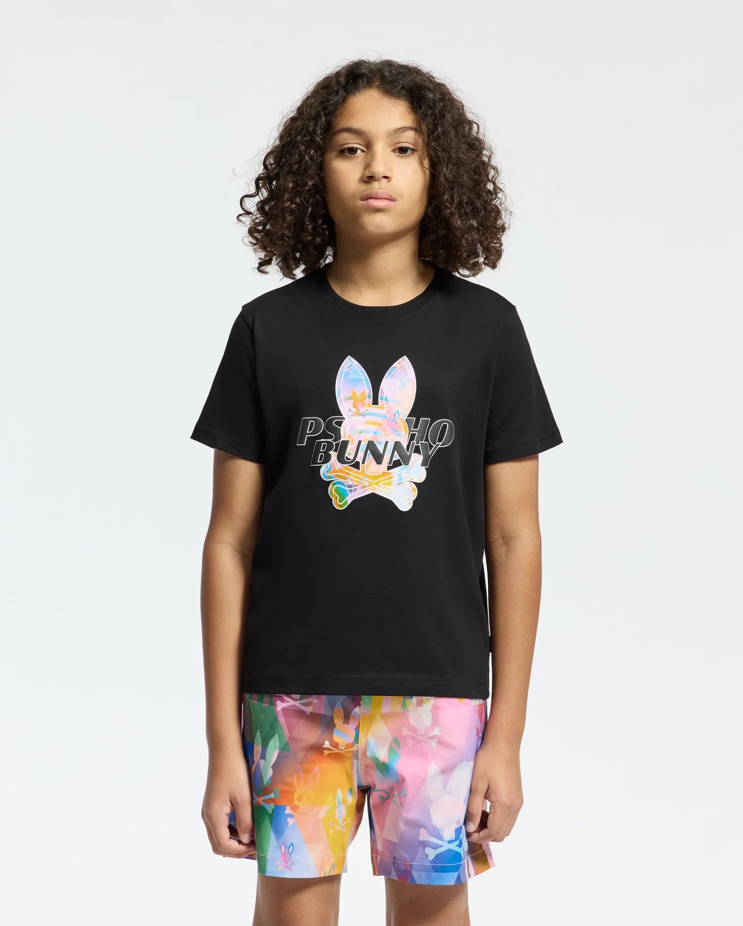 A person with curly hair is wearing a black Psycho Bunny KIDS MILOS GRAPHIC TEE made from Peruvian Pima cotton, with a space-age bunny ears graphic and "BUNNY" text. They're also in colorful abstract pattern shorts on a white background.