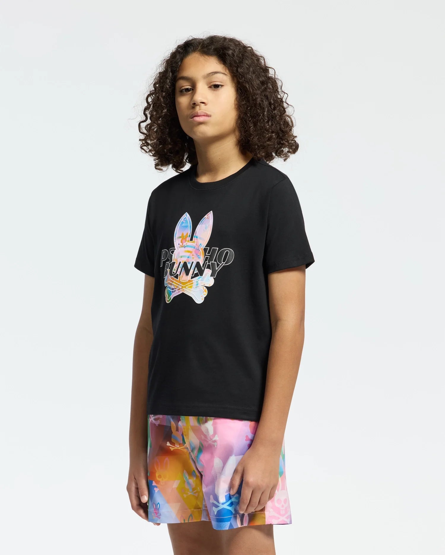 A young person with curly hair wears a black KIDS MILOS GRAPHIC TEE by Psycho Bunny, featuring a Center Bunny Graphic and the word "PLAYBOY." They confidently pair it with a vibrant abstract patterned skirt against a plain white background.